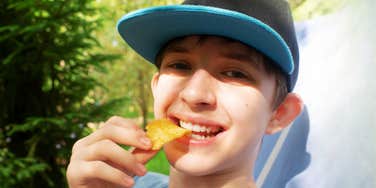 close-up lifestyle shot of a happy pre-teen boy eating snacks outdoors in a green space; illustrating 'the free-range autonomy deficit' where standard, unscripted childhood leisure is replaced by micro-managed scheduling.