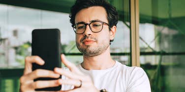 A young Millennial man in a white shirt and glasses smiling as he takes a selfie with his smartphone; a visual representation of 'the performed identity' and the digital social experiment that backfired.