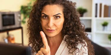 A female worker gesturing a warning from her office desk surrounded by shelves and plants; illustrating the 'social contract breakdown' where veteran employees warn younger generations against blind institutional loyalty.