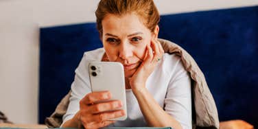 A woman lying in bed pulled under a heavy blanket, focusing intently on her glowing smartphone screen; a visual representation of 'the cloistered consumer' and the reality of millennial screen addiction.