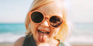 A young toddler girl playfully sticking out her tongue while posing in large sunglasses; illustrating the 'unshakeable self-esteem' and secure emotional foundation given to daughters by supportive, values-driven parents.