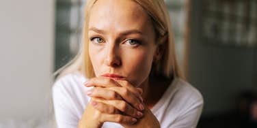 A close-up portrait of a thoughtful blonde woman sitting alone in a living room, looking seriously into the camera with her hands resting on her chin; a visual representation of 'the unveiled grievance' and the hidden reality of maternal regret.