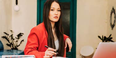 A close-up portrait of a corporate employee in a red blazer working on a laptop inside a modern workspace; illustrating 'the purchasing power disconnect' and the systemic economic beliefs causing division between managers and staff.