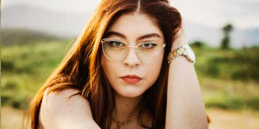 A millennial woman with red hair and glasses focusing intently on the camera in a modern outdoor setting; illustrating the economic pressures and changing family expectations that shape millennial family planning decisions.