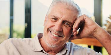 A content senior man smiling peacefully while sitting on a couch in a bright, comfortable living room; a visual representation of 'the affluent realist' enjoying a secure, self-made retirement.