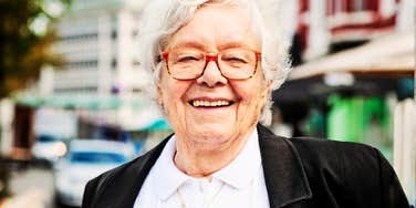 A close-up street portrait of a cheerful, vibrant elderly woman with a soft bokeh background; illustrating 'the vital aging paradigm' and the rejection of standard fears surrounding growing older.