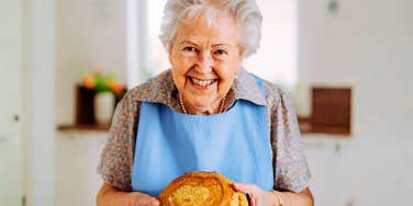 A close-up portrait of a cheerful elderly woman showcasing a homemade rustic bread loaf in a home kitchen; illustrating the 'analog renaissance' and the timeless satisfaction found in scratch-cooking hobbies.