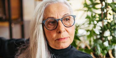 A serious mature woman looking thoughtfully to the side in a softly lit home setting; illustrating a 'cultural protocol shift' and the generational disconnect over traditional manners and respect.