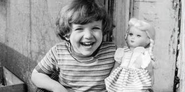 A retro black-and-white style snapshot of a young child playing alone with a toy doll near a balcony railing; illustrating 'the architectural change in play' where historic youth independence contrasts with modern supervised childhoods.