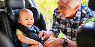 A happy senior man securing a cute toddler boy into a child safety seat for a road trip; illustrating a critical 'parental authority breach' and the breakdown of trust between parents and grandparents.