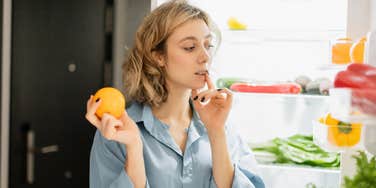 thoughtful woman looking inside fridge holding orange