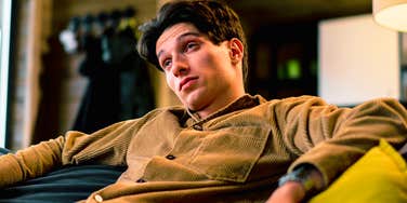A close-up portrait of an exhausted young man on a couch after watching TV, still holding the remote; illustrating 'cognitive buffering' and the subconscious reliance on media to quiet a racing mind at bedtime.