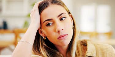 A close-up portrait of an anxious woman resting her chin near her hands in deep thought on a couch; illustrating 'the learned helplessness loop' where repetitive negative self-talk locks an individual in emotional stagnation. 