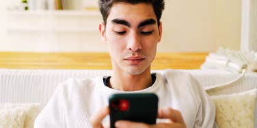 A man sitting indoors, completely absorbed as he types a message on his smartphone; a visual representation of 'textual tension' and the subtle text-ending habits that research says can make a sender come across as rude.