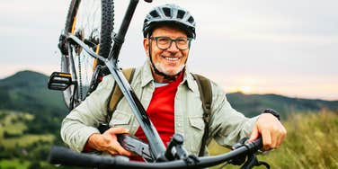 Happy older man carrying his bicycle through nature, reflecting how staying active and doing challenging things helps people enjoy life at any age