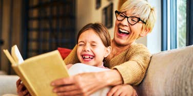 A grandma sharing a humorous moment with her grandchild over a children's storybook; illustrating 'cognitive vitality' and the spontaneous, late-life emotional healing found in intergenerational play.