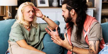 A close-up lifestyle shot of a young couple or roommates sitting in a tense posture in a home flat; illustrating 'the cognitive dissonance standoff' where a lack of internal insight leads to external blame-shifting during a dispute.