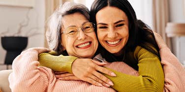 An older mom wearing glasses widely smiling while her daughter hugs her from behind in a home setting; illustrating the 'generational disconnect' and the 'me-first' behavioral patterns reported by modern parents.