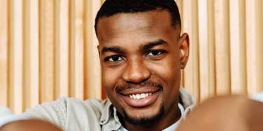 A casually dressed African American man sitting indoors and smiling warmly; a visual representation of 'the protective softness' and the subtle signs a man loves you deeply without saying it.
