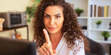 A close-up portrait of a corporate employee giving a firm warning gesture from her modern desk with plants and shelves behind her; illustrating 'the intellectual guardrail' model where setting limits signals high cognitive capability.