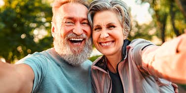 A close-up portrait of an attractive gray-haired man with a beard and an elegant senior woman smiling while capturing a digital memory; illustrating 'active longevity' and the daily routines that reflect deep internal fulfillment.