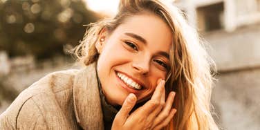 A candid, radiant close-up of a happy young woman smiling warmly in a blurry open-air environment; illustrating the 'unobserved bliss' model where true life enjoyment manifests in moments of quiet solitude.