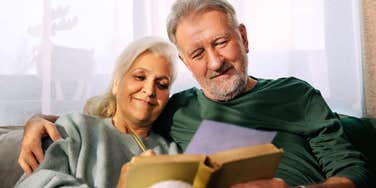 A young couple sitting peacefully together on a couch, focused on reading a book in a softly lit room; a visual representation of 'the parallel play ideal' and the foundational marriage habits older generations mastered.