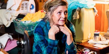 A senior woman sitting on a chair in a heavily cluttered room filled with stacked boxes, piles of clothing, and miscellaneous household items; a visual representation of the 'overwhelmed resident' archetype used to study the psychology of chronic disorganization.