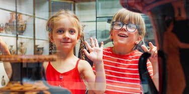 A close-up portrait of two young kids looking eagerly through a clear display window inside a building; illustrating 'the institutional isolation syndrome' where physical safeguards limit tactile exploration.