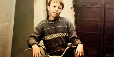 A vintage, faded analog photograph from the 1970s of a young boy riding a retro bicycle in a grassy yard, with a classic mid-century car parked in the background; a visual representation of 'the autonomous child' archetype used to study Gen-X latchkey independence.
