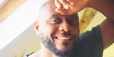 An indoor lifestyle portrait of a relaxed, happy man looking through a window with a gentle, loving smile, embodying patience, emotional intelligence, and relational satisfaction.