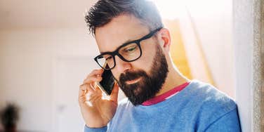 Front view of a man looking down with a tense expression while holding a smartphone in a room; illustrating 'the cognitive load of deception' archetype where body language leaks internal conflict.