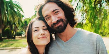 Close-up profile of a happy husband and wife sharing a spontaneous laugh outdoors against a soft-focus background of green trees; illustrating 'the relational resonance model' where long-term partners mirror each other's joy.