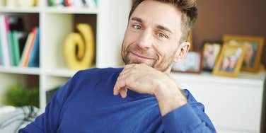 A gentle, smiling man sitting on a couch in a brightly lit room, capturing the 'psychological safety' and baseline warmth found in thriving, long-term relationships.