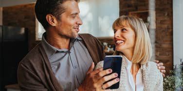 A relaxed husband and wife sitting together on a modern couch, smiling and laughing while looking at a smartphone screen; a visual representation of 'the connected leisure' archetype used to study long-term marital friendship.