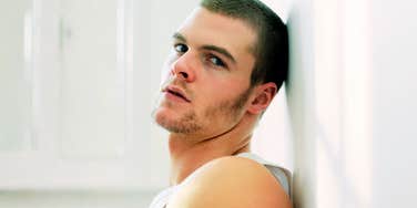 A man leaning against a plain wall with a detached expression and crossed arms, illustrating emotional distance, passive disrespect, and relational disengagement.