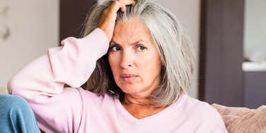 A pensive middle-aged woman in a cardigan rests her head on her hand while sitting on a living room couch; illustrating 'the midlife identity metamorphosis' where deep psychological restructuring occurs.