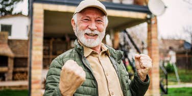 A contented senior man with gray hair sits comfortably on a wooden deck, smiling gently while holding a cup of coffee. He looks out over a calm, expansive view of mountains and rolling hills during golden hour lighting.