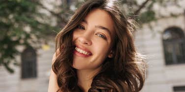 A close-up portrait of a serene young woman smiling softly while looking into the camera, with a blurred cityscape in the background conveying inner calm and emotional stability.