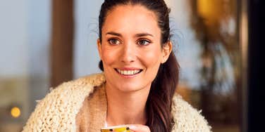 A smiling brunette woman sitting outdoors with her friend on a crisp autumn day, holding a warm cup of tea while enjoying a captivating conversation.