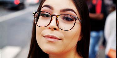 A close-up portrait of a thoughtful young woman wearing tortoiseshell glasses, a blue top, and a light trench coat with a patterned scarf, looking at camera with an observant, analytical expression on a city street.