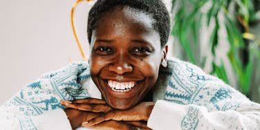 A close-up lifestyle portrait of a happy African woman smiling warmly at the camera, wearing a soft patterned knit sweater inside a brightly lit modern apartment, exuding comfort and emotional intelligence.