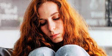 Indoor apartment portrait of a sad redhead woman curled up on a couch, looking down thoughtfully with a worried expression; illustrating the physical and mental fatigue of deep emotional processing.