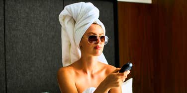 relaxed female hotel guest with her hair wrapped in a white bath towel, comfortably using a remote control from a plush mattress, highlighting premium hospitality standards.