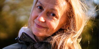 A close-up portrait of a beautiful mature woman with silver-streaked hair smiling serenely outdoors in golden autumn sunlight; a visual representation of 'the grounded elder' archetype used to study aging with dignity.