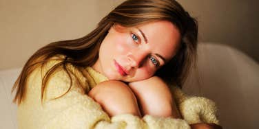 A close-up portrait of a sad blonde woman aged 25 to 30 wearing a knitted yellow sweater, sitting on a couch and holding her legs while looking directly at the camera; a visual representation of 'the guarded self' archetype used to study behavioral habits of emotional insecurity.