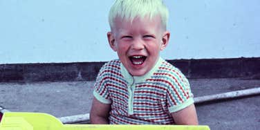 A vintage 1970s film photograph of a young boy in a white t-shirt and jeans pushing a large yellow toy dump truck down a garden path; a visual representation of 'the carefree epoch' used to illustrate childhood resilience.