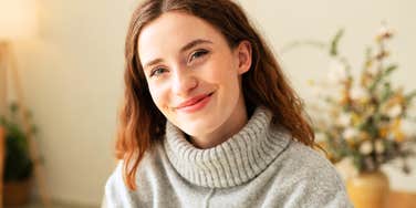 Cute young redhead woman with an engaging grin sitting perched on the end of a table looking at the camera with smiling eyes.