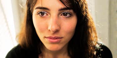 A close-up portrait of a woman with a worried expression, her hands raised defensively to her head in a dark environment; a visual representation of 'the internal critic' archetype used to study cognitive distortions in low self-esteem.