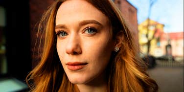 A portrait of a confident young woman with curly red hair and a black blazer standing outdoors in front of a contemporary glass office building; a visual representation of 'the guarded independent' archetype used to study defensive detachment in modern dating.